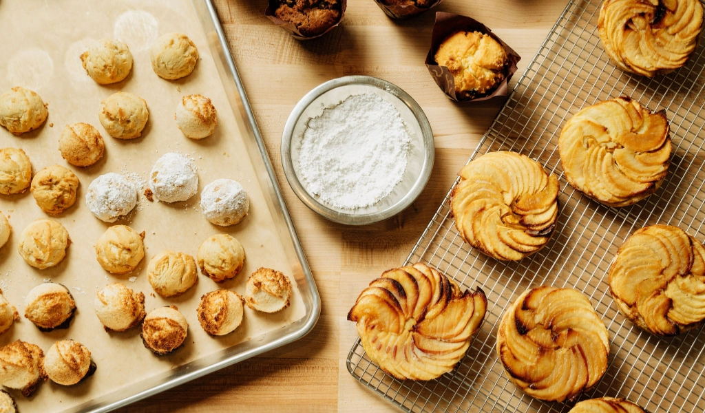 A baked goods assortment featuring powdered sugar cookies on a baking sheet, apple pastries on a cooling rack, and muffins, all centered around a bowl of powdered sugar.