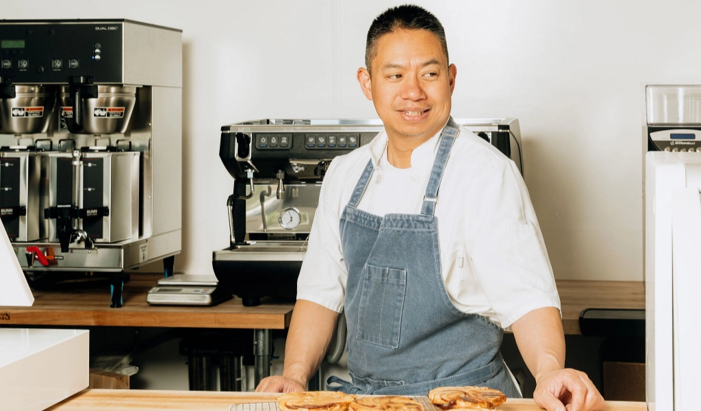 A chef in a blue apron stands behind a wooden counter with pastries at Preligen Artisan Bakery.