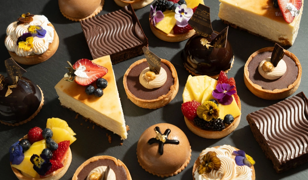 An assortment of various desserts from Preligen Artisan Bakery, including cheesecake slices, chocolate tarts, fruit tarts, and other decorated pastries displayed on a dark surface.