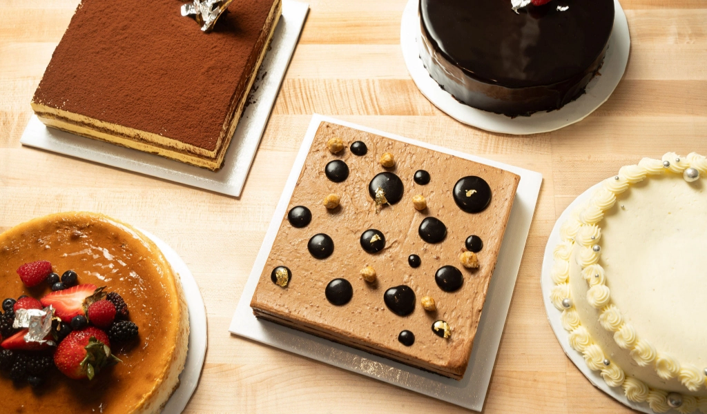 An assortment of five decorated cakes placed on a wooden surface, featuring a variety of flavors and toppings including fruit, chocolate glaze, and decorative elements.