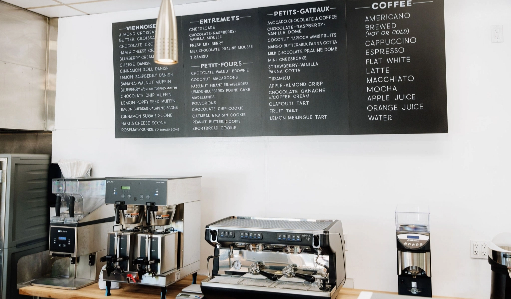 The cafe counter at Preligen Artisan Bakery with coffee machines, a grinder, and a menu board listing various high-end French pastries, petit-fours, and coffee options.