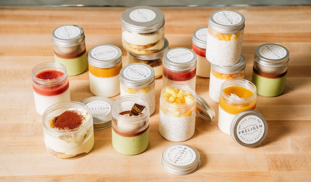 An assortment of high-end French pastries in small glass jars with metal lids, arranged on a wooden surface at a luxury cafe.