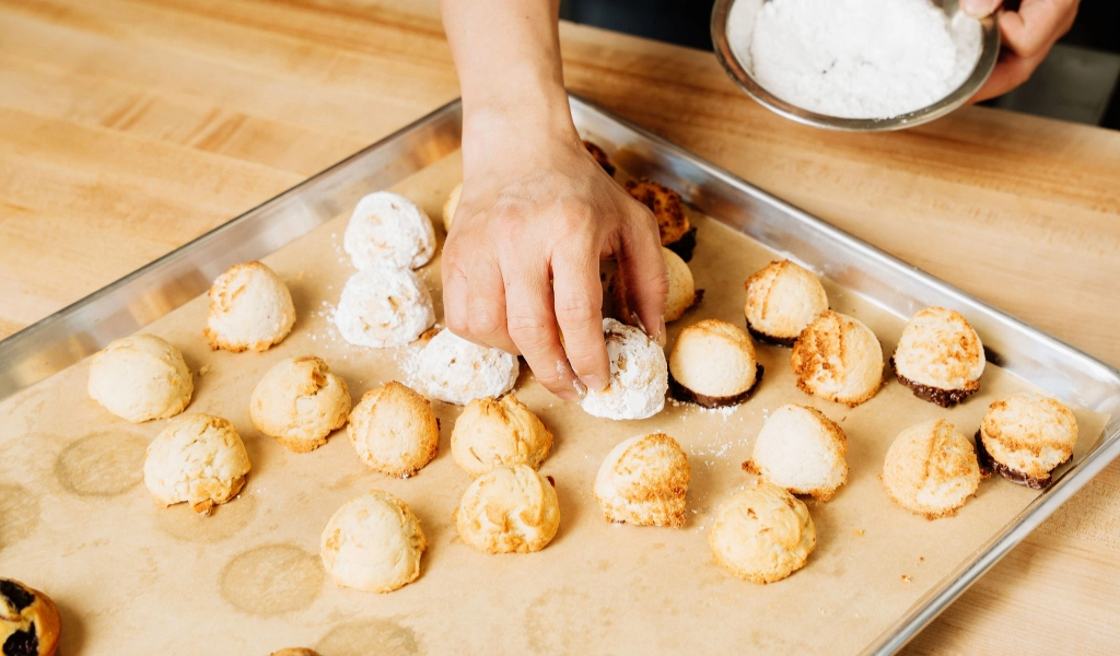 A person at Preligen Artisan Bakery is delicately dusting powdered sugar on small round cookies arranged on a baking tray lined with parchment paper.