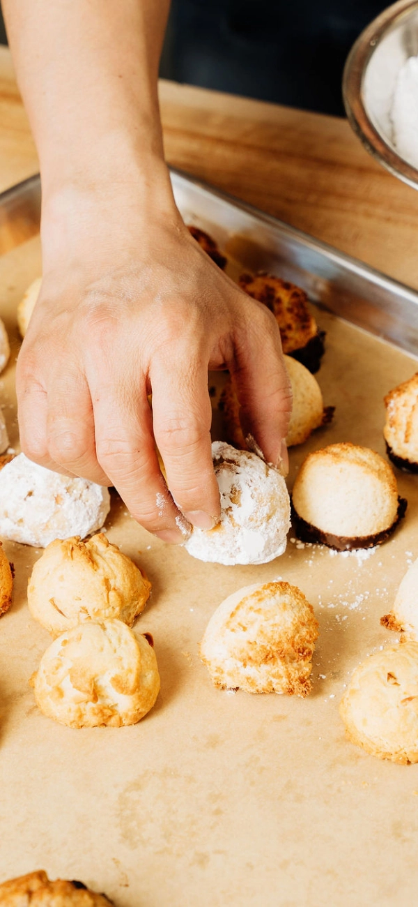 A hand places a powdered sugar-covered cookie among other golden-brown treats on a parchment-lined tray.