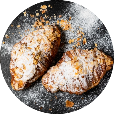 Two almond croissants dusted with powdered sugar are placed on a dark, textured surface.