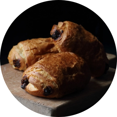 Three chocolate croissants stacked on a wooden board against a dark background, showcasing the exquisite offerings from our high-end French pastries at San Jose bakery.