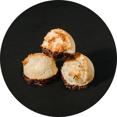 Three coconut macaroons partially dipped in chocolate are stacked against a black background, showcasing the exquisite craftsmanship of our Artisan Bakery.