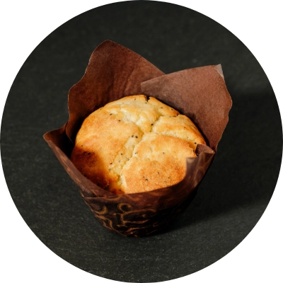 A lemon flavored muffin with poppy seeds wrapped in brown parchment paper on a dark surface.