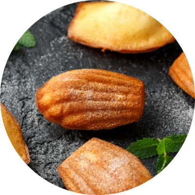 A close-up of golden-brown madeleine cookies on a dark surface, garnished with a few green mint leaves.