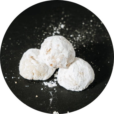 Three powdered sugar-coated cookies are stacked on a dark surface, with additional powdered sugar sprinkled around them.