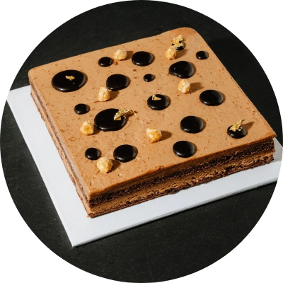 A square layered praline cake with glossy dark chocolate drops on top, garnished with small edible gold flakes, presented on a white platter.