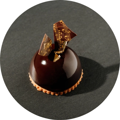 A dark chocolate dome dessert from Preligen Artisan Bakery, showcased on a black surface, topped with decorative pieces featuring musical notes.