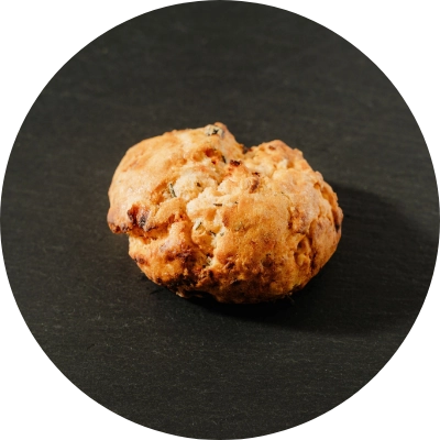 A single scone from Preligen Artisan Bakery sits on a dark surface, featuring a golden-brown crust.