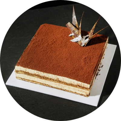 A square tiramisu cake from a San Jose bakery, topped with cocoa powder and decorative chocolate pieces, is elegantly placed on a white square plate against a dark background.