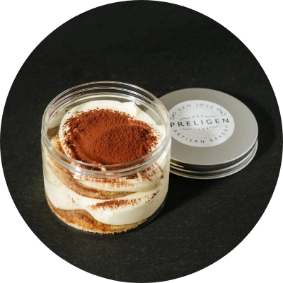 A jar of tiramisu topped with cocoa powder sits next to its lid on a black surface, showcasing the craftsmanship of Preligen Artisan Bakery.